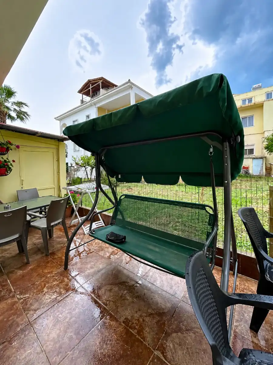 Shaded terrace at Maria Apartments with a garden swing seat, table and chairs beside potted flowers