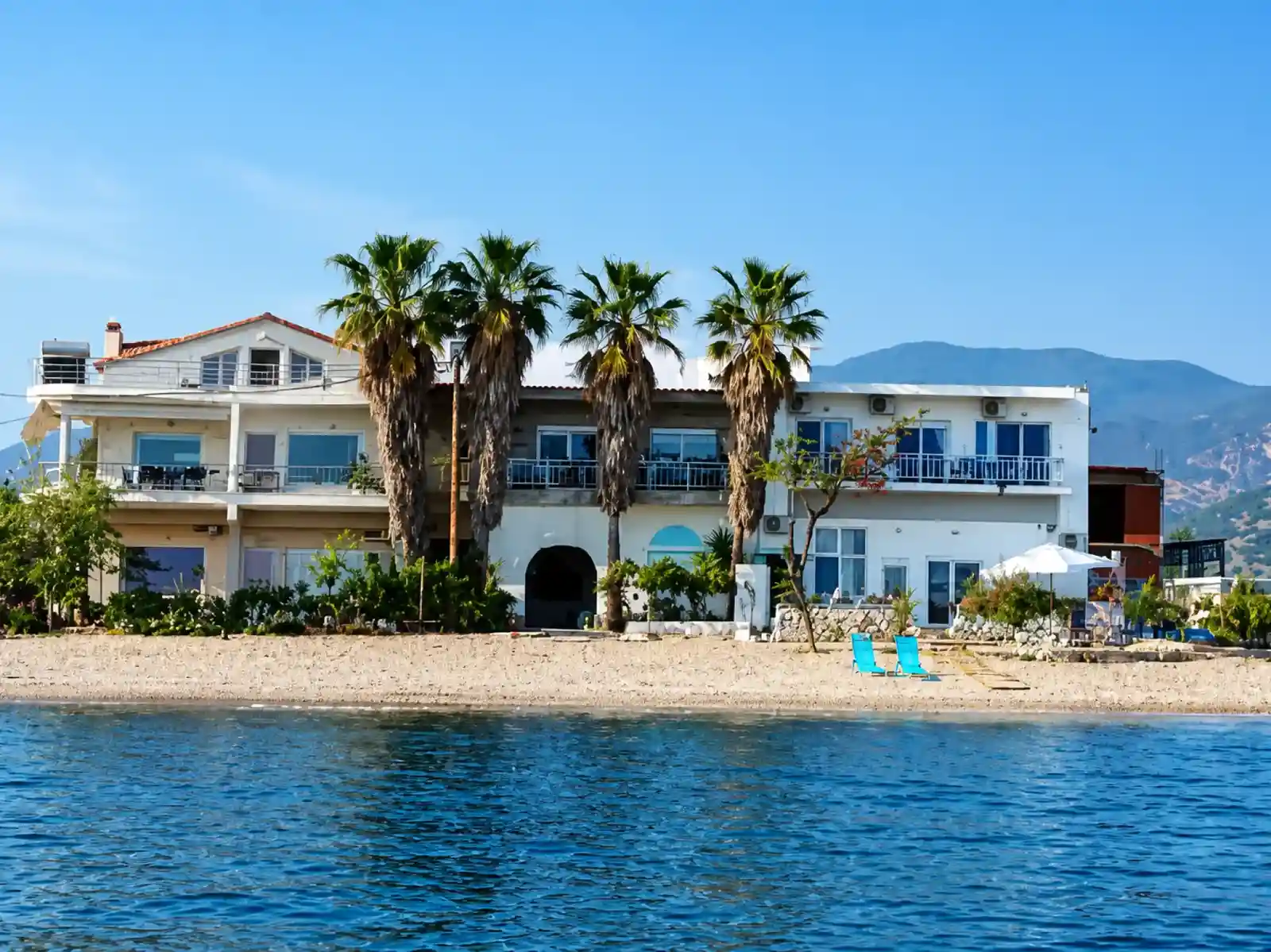 Maria Apartments seen from the water, a white building behind palm trees on the beach in Néa Karváli