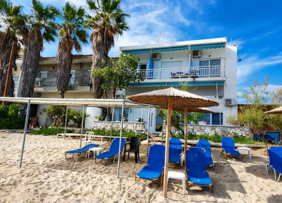 Maria Apartments behind palm trees, with blue sun loungers and a straw parasol on the sand in front